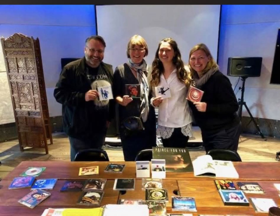 Katiana Giacona (second from right) with friends at a Talisman Lifestyle event, holding up Record Plant album covers over a table spread with vinyl and CDs from the studio's discography.
