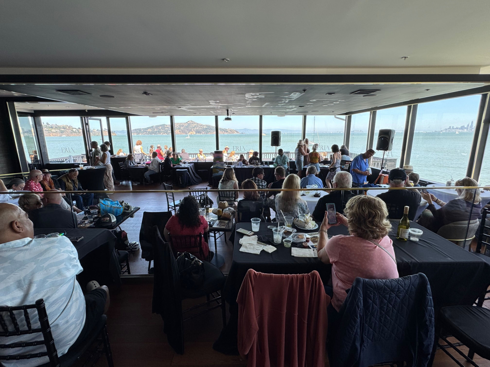 Crowd seated at black-draped tables inside ERIA, the ERIA lettering etched on the window glass and Belvedere, Angel Island, and the bay beyond