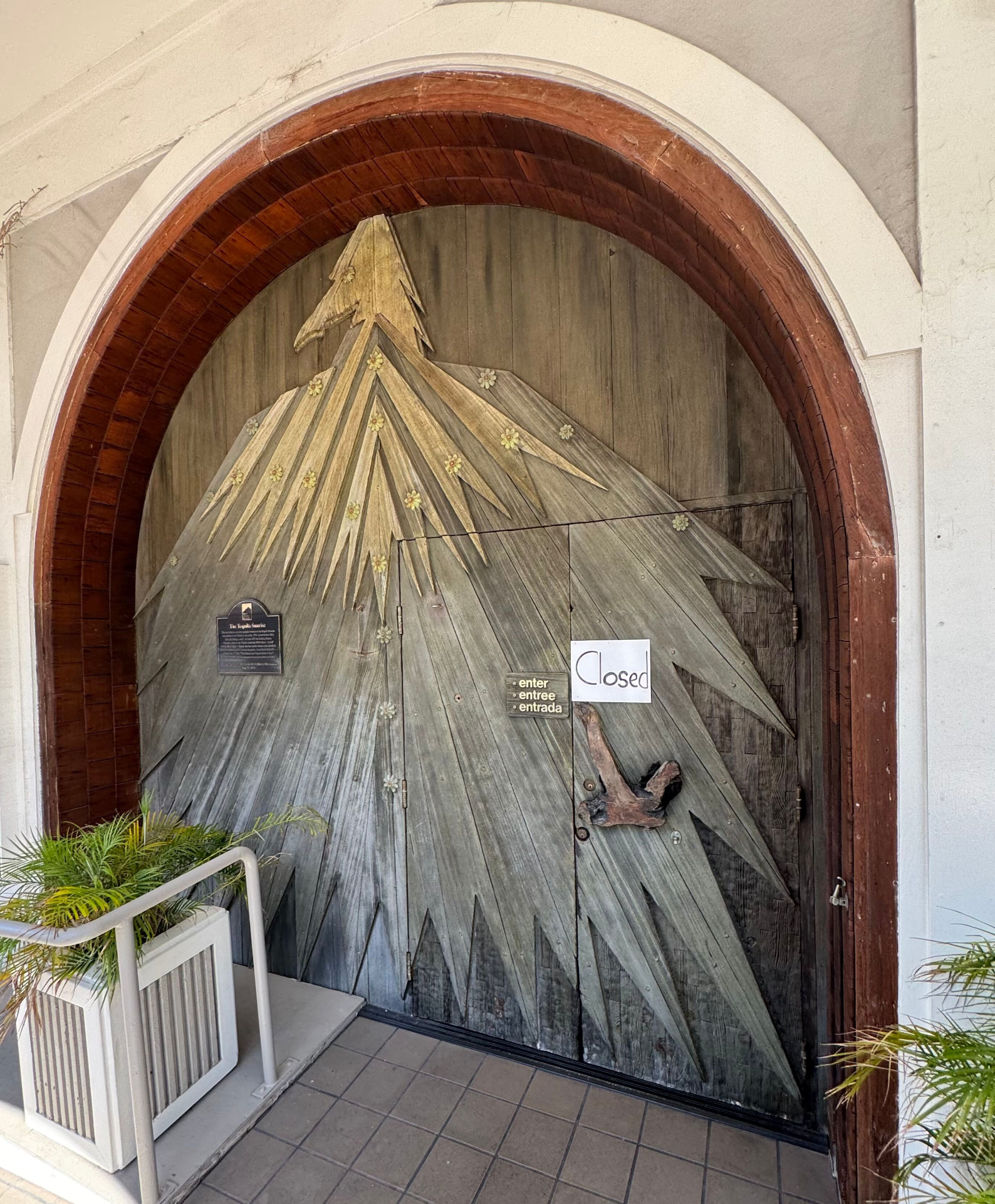 The Trident's carved wooden sunburst doors inside a redwood archway, the Tequila Sunrise bronze marker bolted to the left panel and a handwritten Closed sign taped to the right