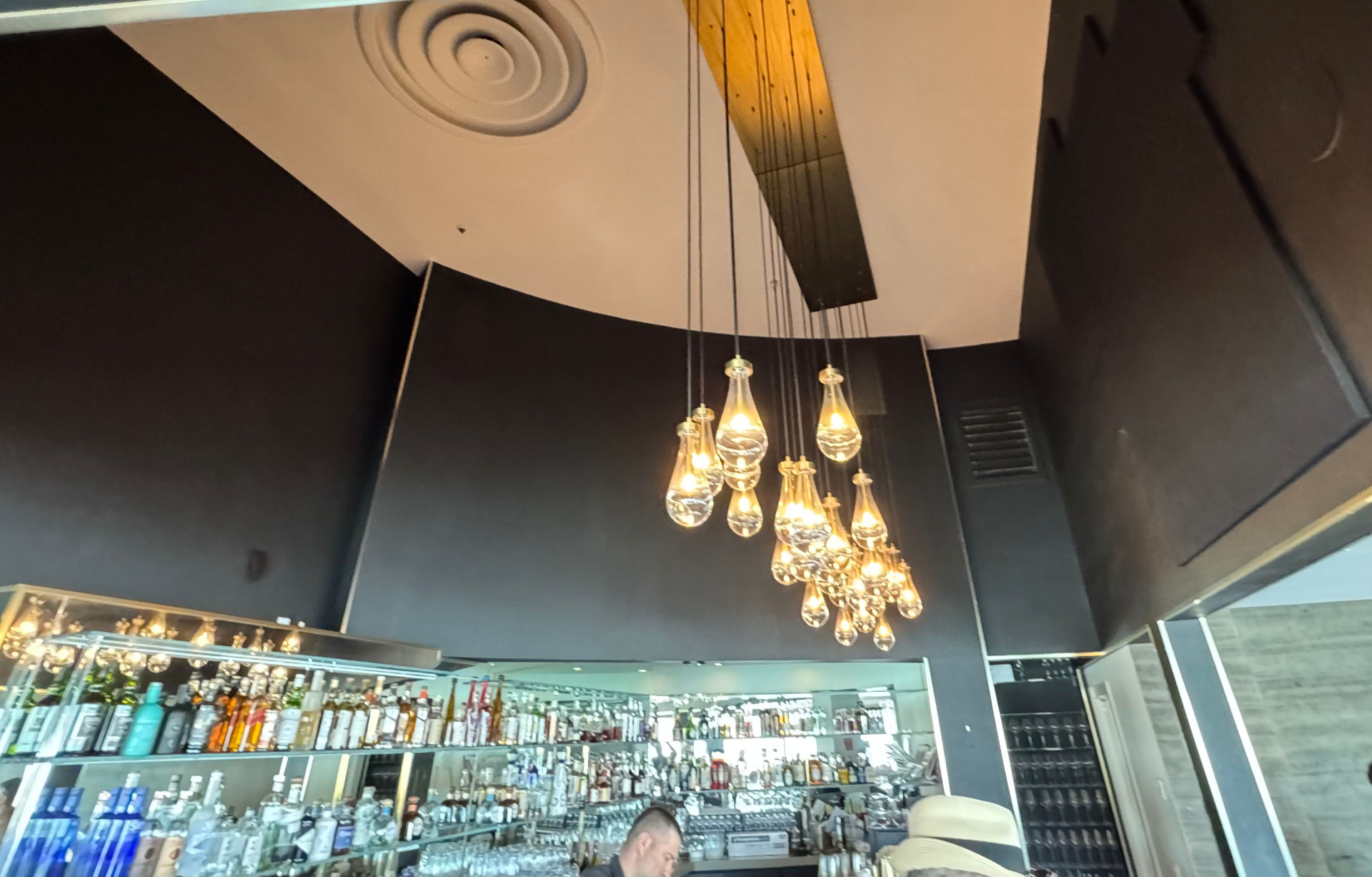 Carved concentric spiral medallion and bare wood beam above the dark soffit at ERIA, glass teardrop pendants hanging over the backlit bar below