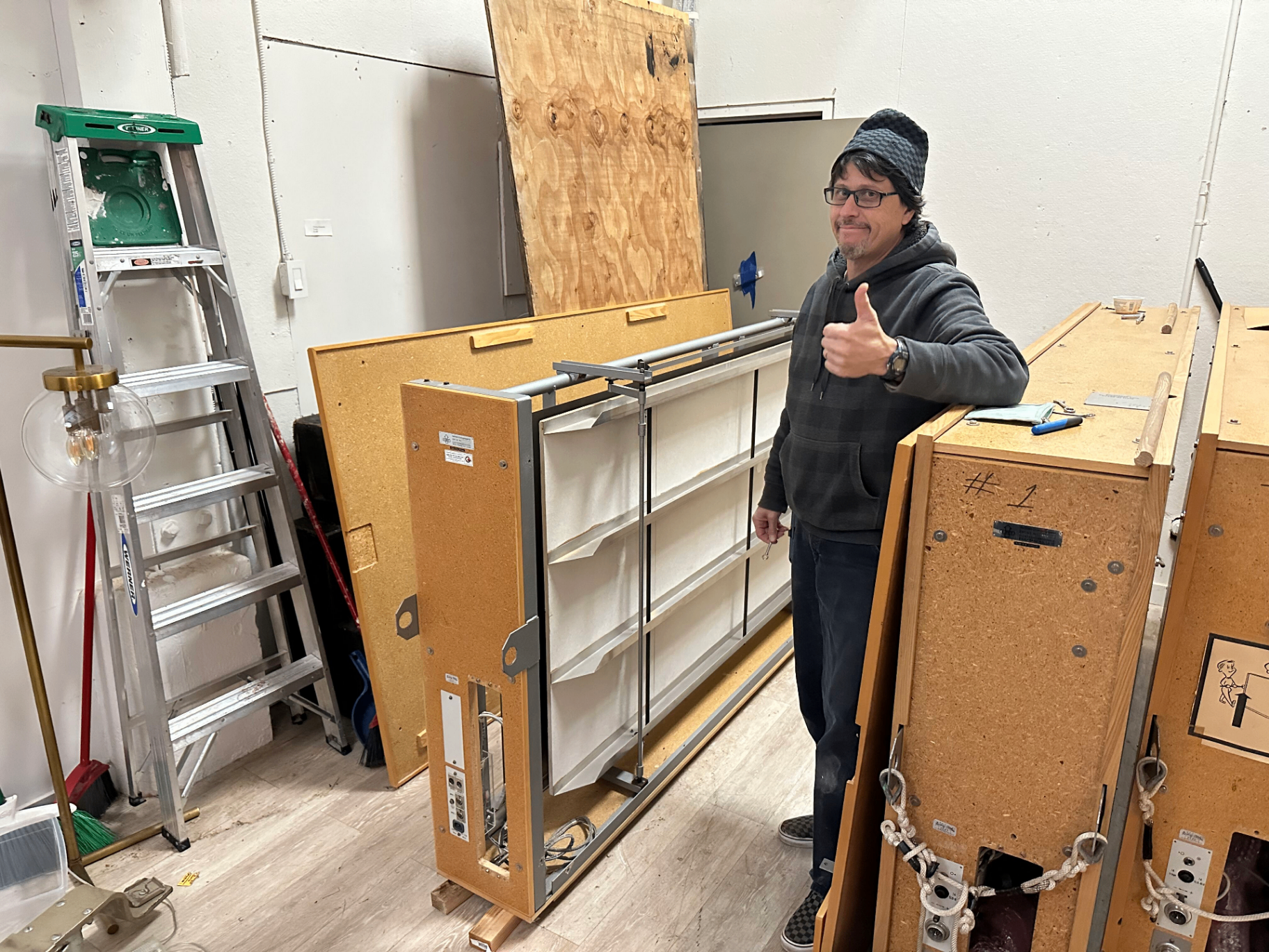 A specialized technician in a beanie and hoodie giving a thumbs up next to a disassembled plate reverb unit in the back area of 2200 Studios, with a ladder, plywood, and other plate units visible around the workspace.