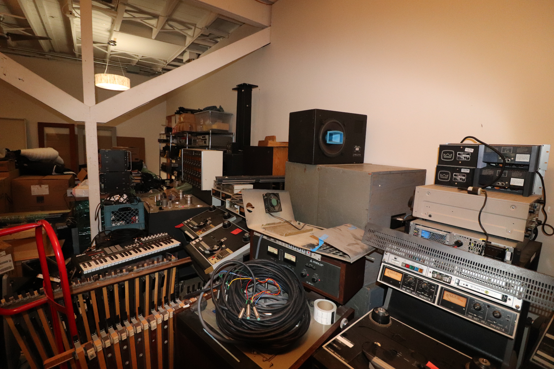 Back area of 2200 Studios stacked with vintage outboard gear, speakers, keyboards, cables, and road cases under a vaulted ceiling.