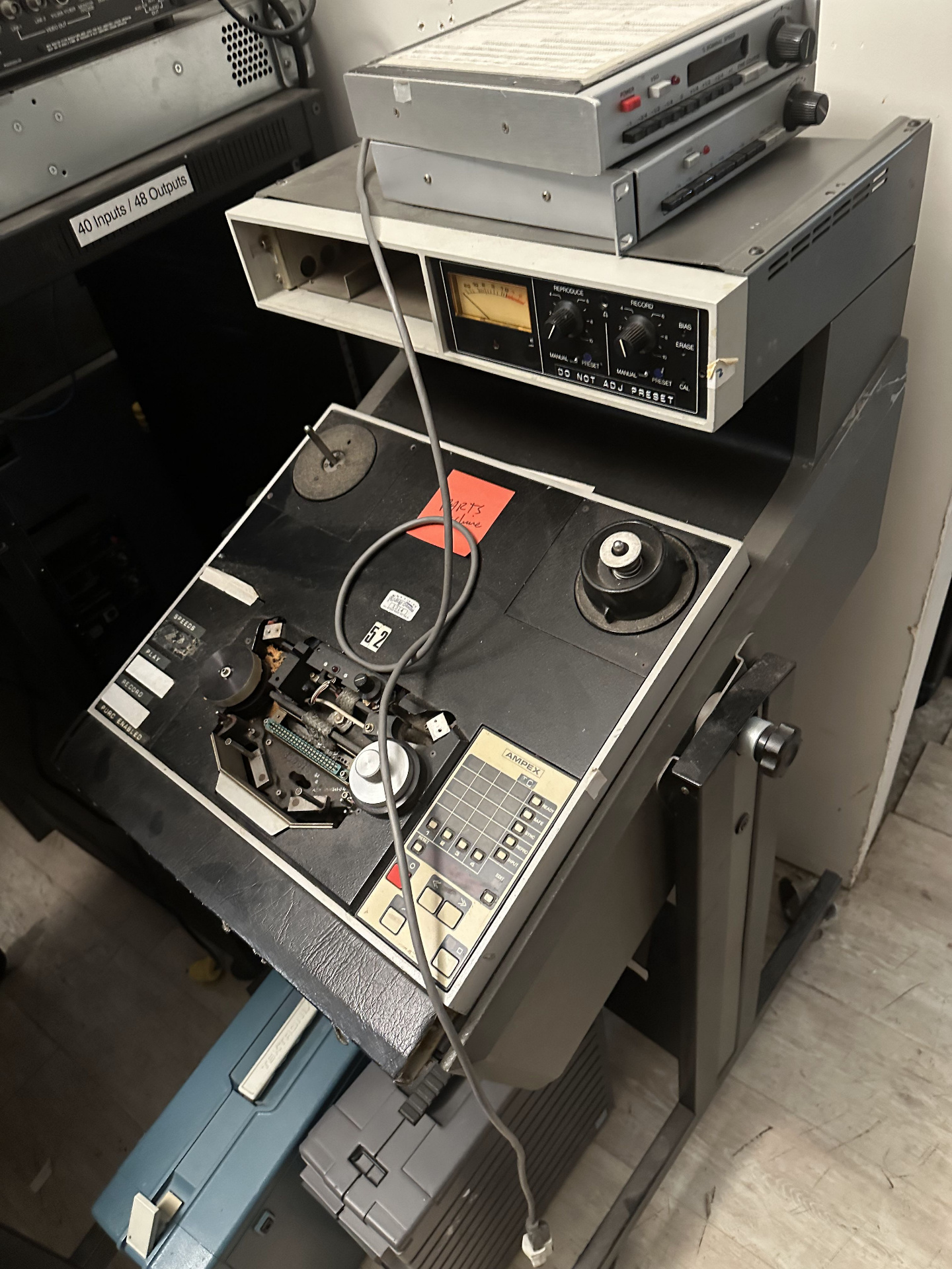 Vintage Ampex multitrack tape machine with exposed transport mechanism, sitting beside racks labeled 40 inputs / 48 outputs in the back area.