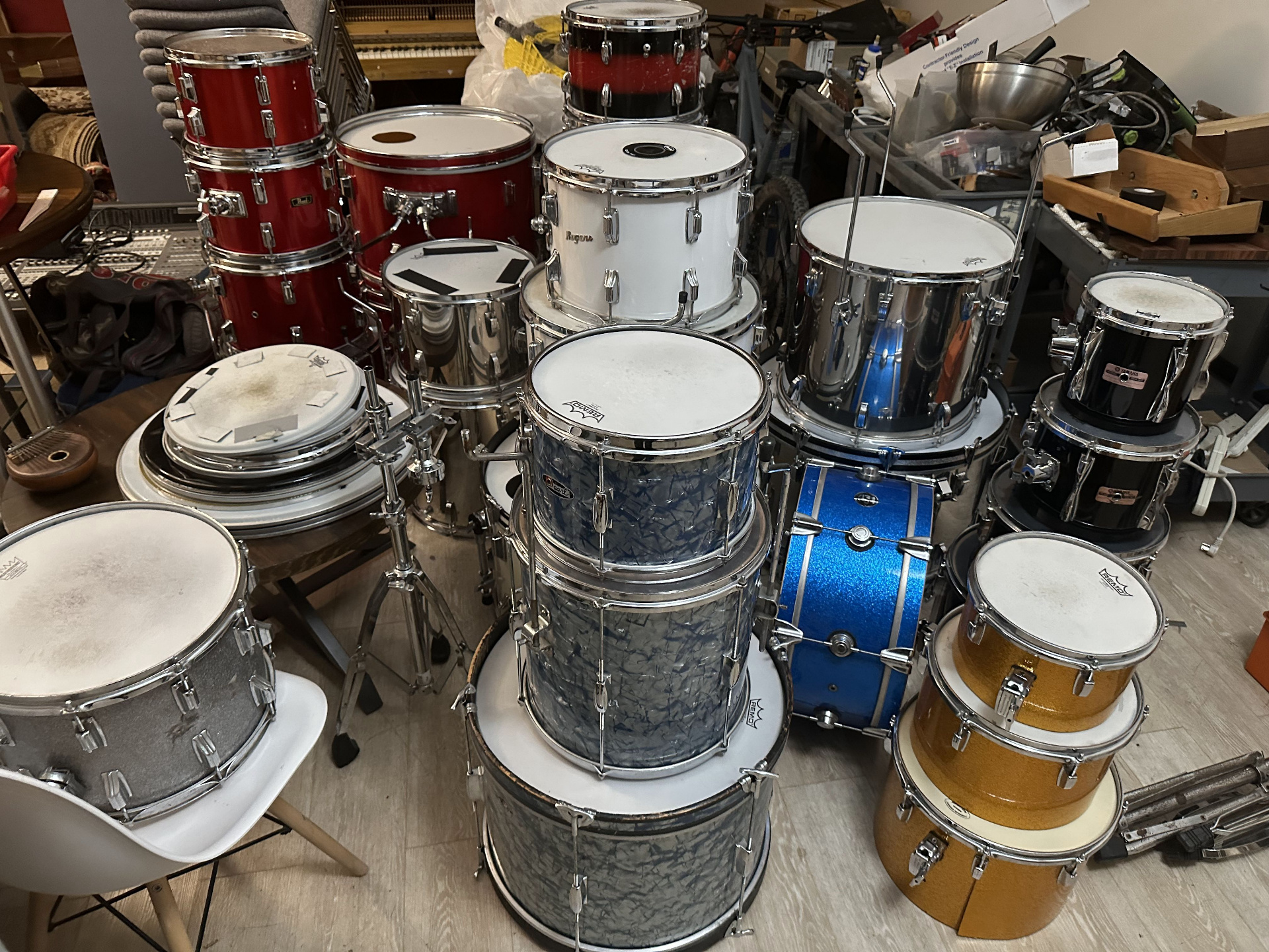 A dense collection of vintage drum kits stacked in the back area — red, white, blue sparkle, black pearl, and gold finishes piled together.