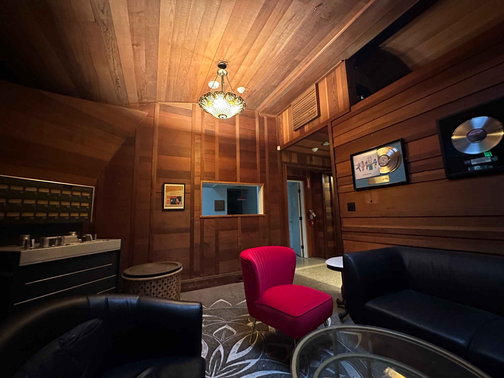 The game room in 2026 — redwood-paneled walls, a red velvet chair, black leather seating, vintage chandelier, and framed gold and platinum records.