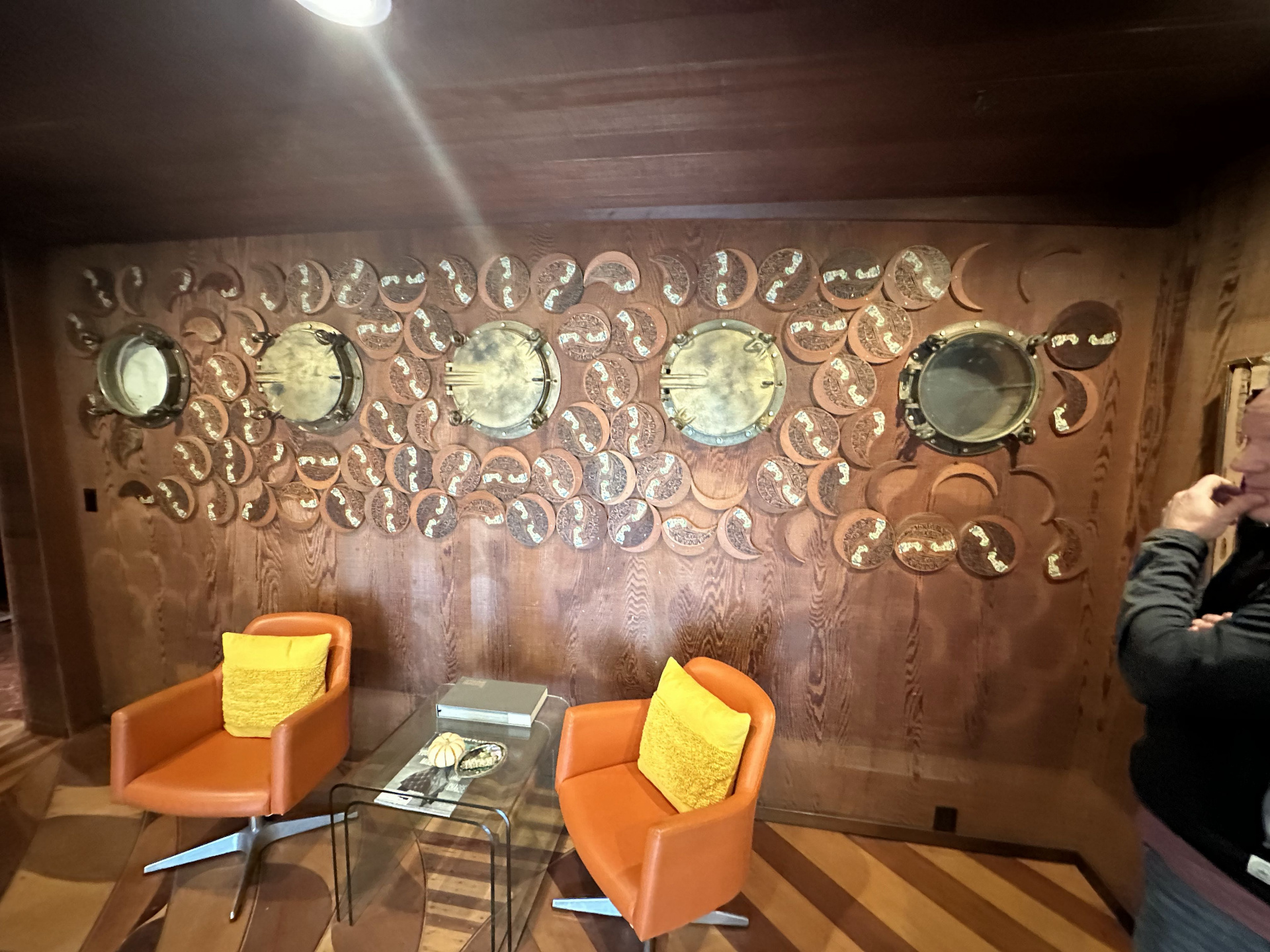 Carved redwood lobby wall set with five brass ship portholes and dozens of carved moon and crescent panels, with two orange leather chairs in the foreground.