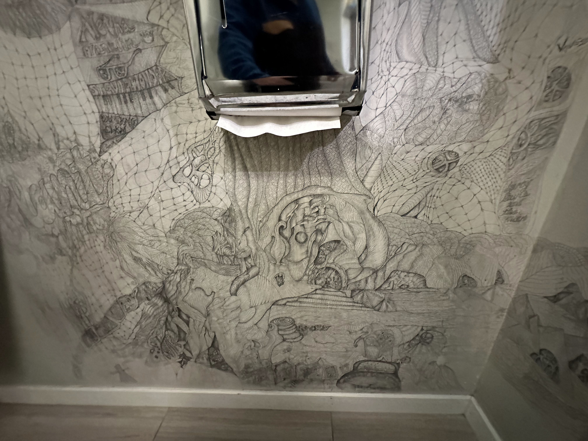 Another section of the bathroom pencil mural — dense, intricate line work spreading across the wall around a paper towel dispenser.