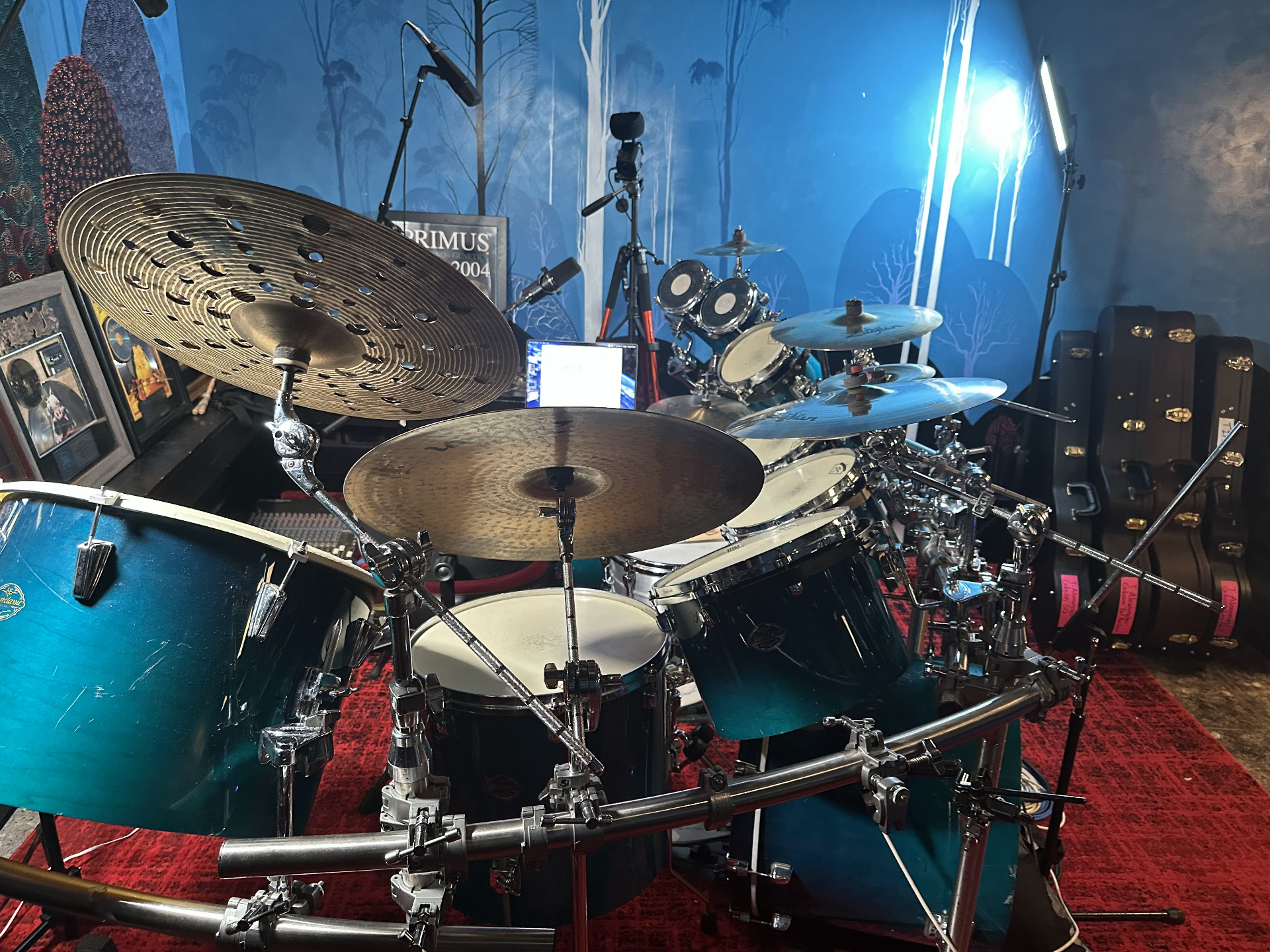 A teal-finish drum kit with a large perforated ride cymbal set up on a red rug, with a framed Primus Animals Should Not Try to Act Like Centaurs award in the background.