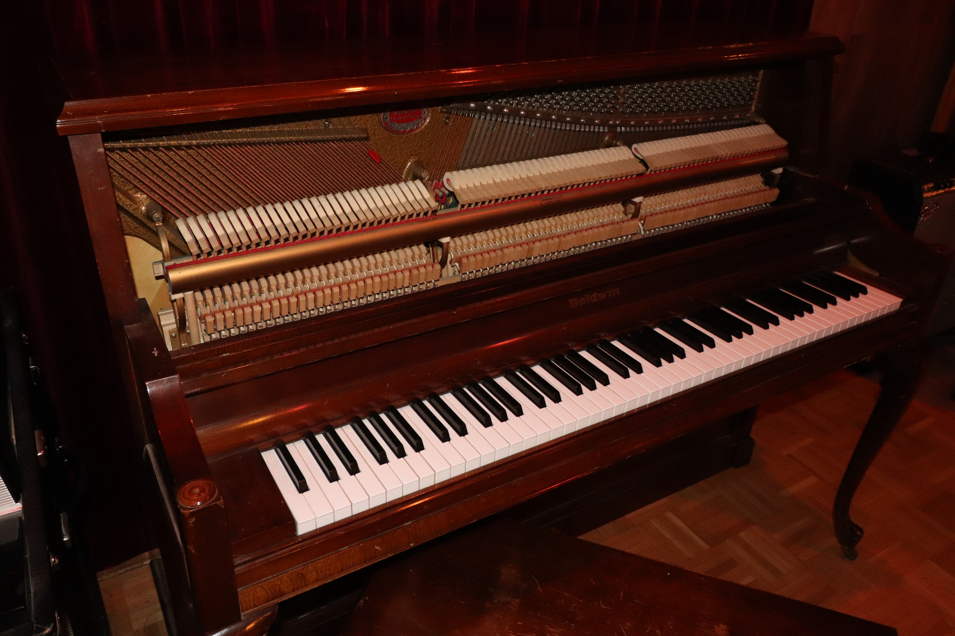 Baldwin upright piano in Studio B with the front panel removed, exposing the hammers, strings, and tuning pins, set against deep red drapery and parquet floor.