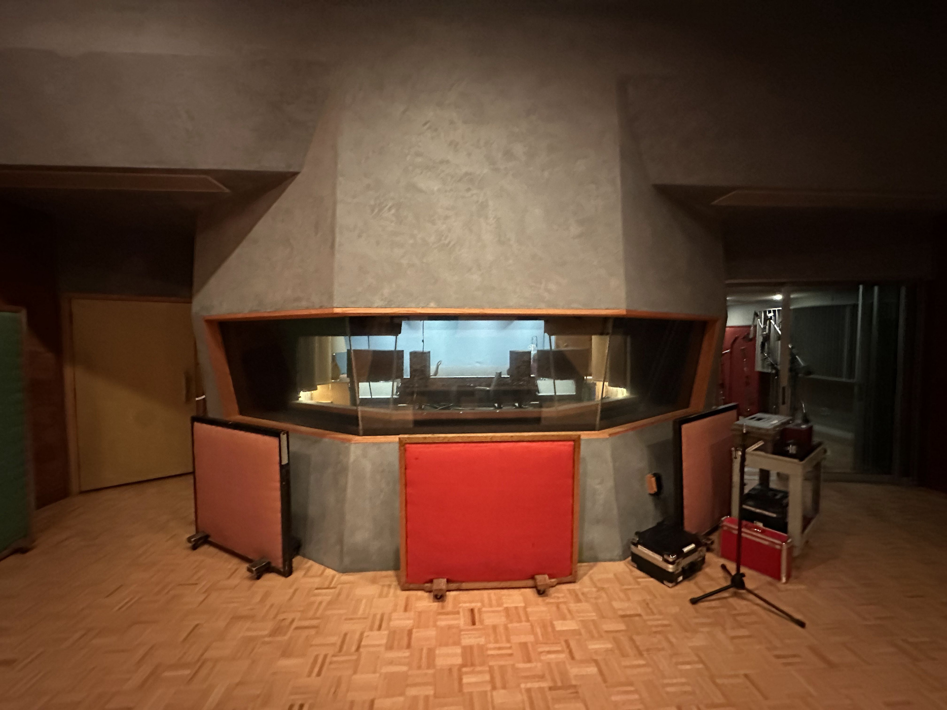 The angled control room window of Studio A viewed from the live room — orange-paneled console base set into a textured grey wall, parquet floor in the foreground.