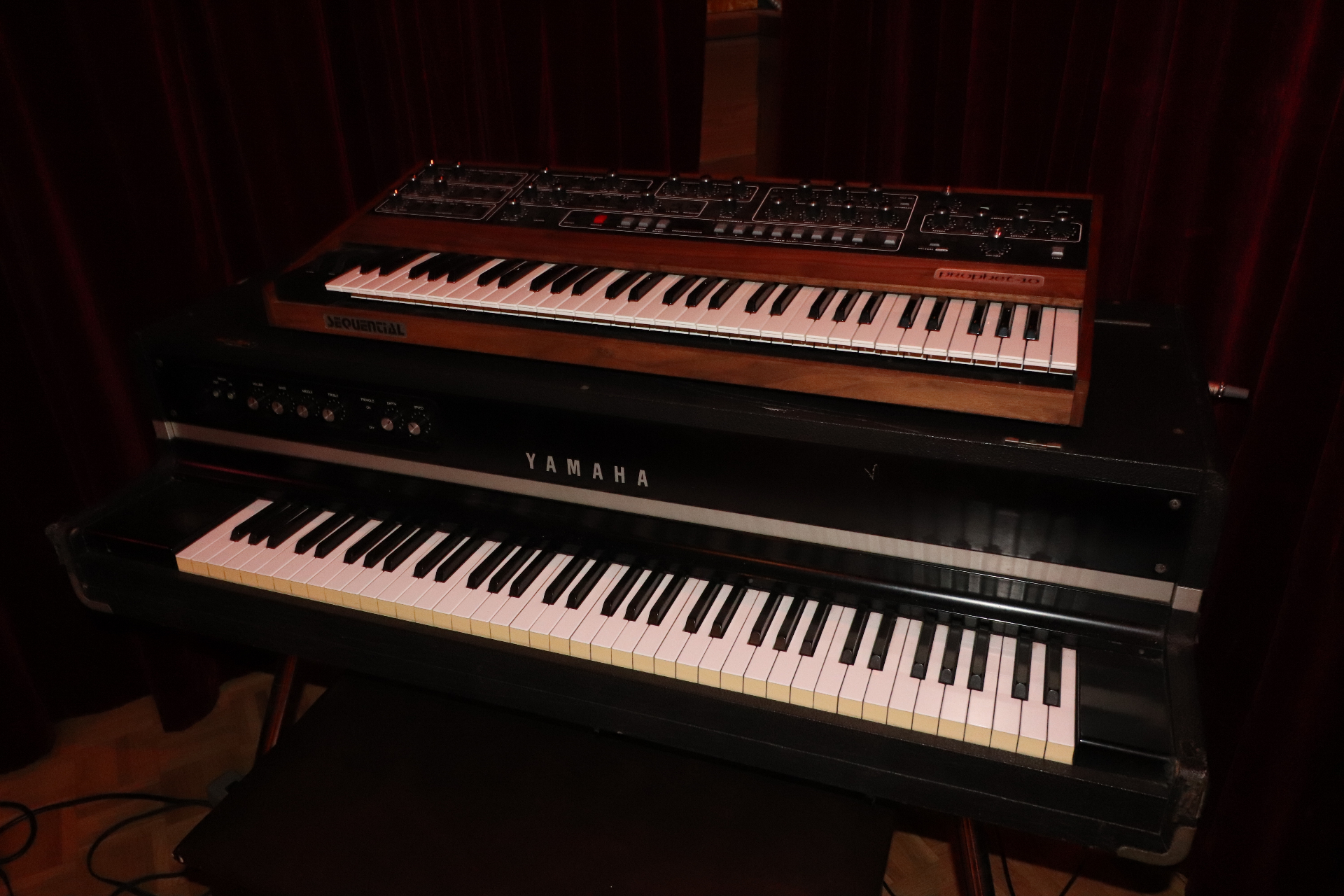 A Sequential Prophet-10 analog synthesizer stacked on top of a Yamaha electric piano in Studio B, set against deep red velvet curtains.