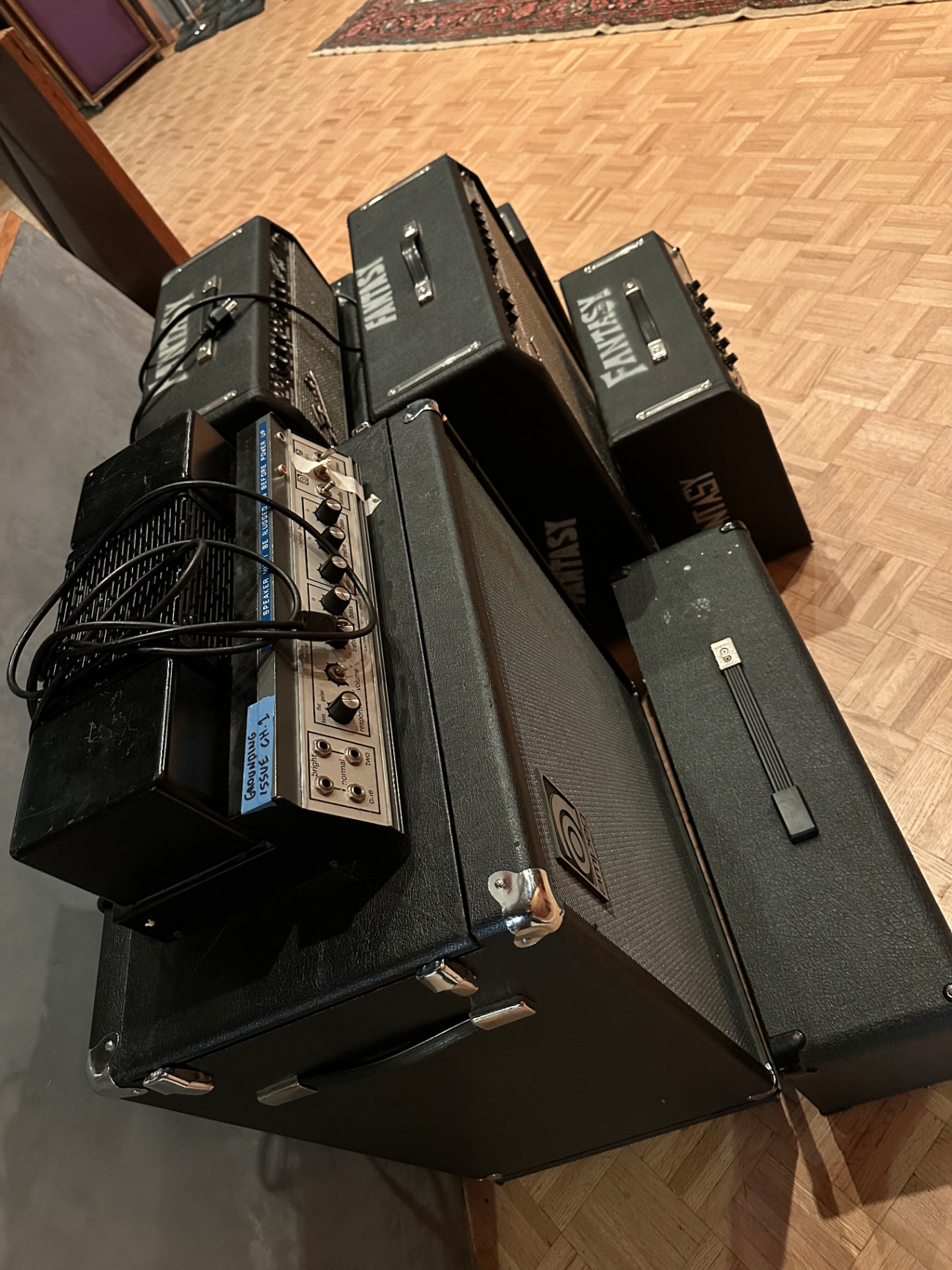 A cluster of vintage Fender and Ampeg amplifiers stenciled 'FANTASY' stacked on a parquet floor, with a tube head and patch cables on top.