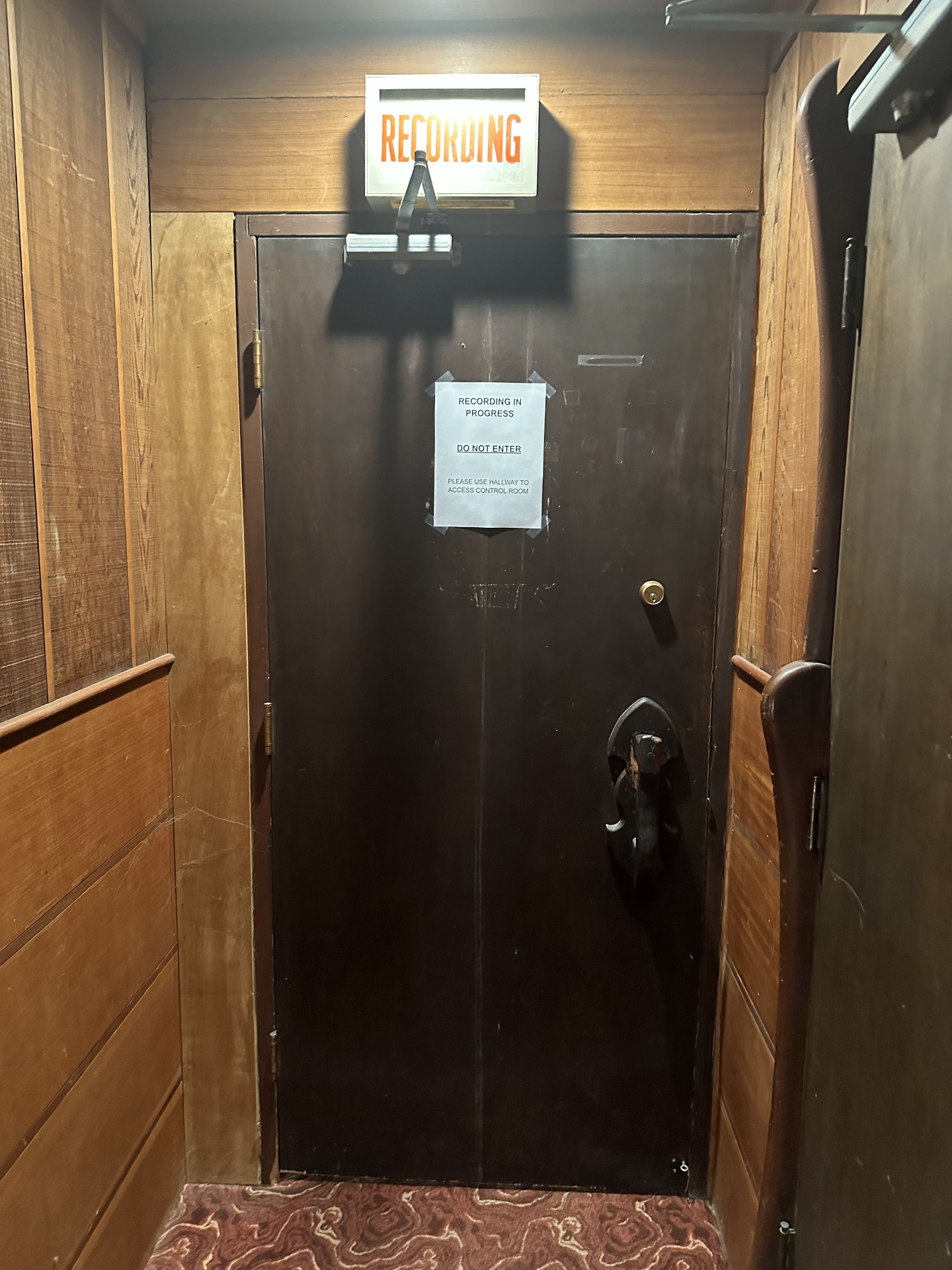 The Studio A door — orange 'RECORDING' light glowing above a dark wood door with a taped 'Recording in Progress — Do Not Enter' sign, set into redwood paneling above the maroon swirl carpet.