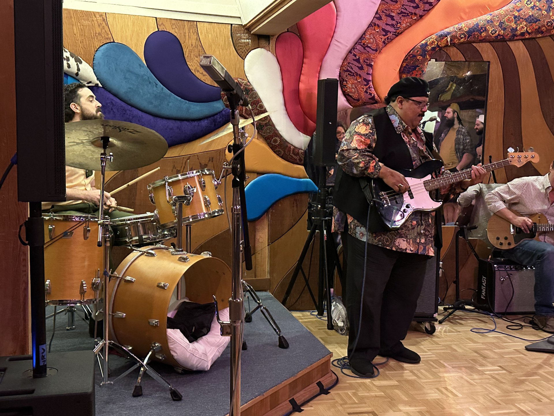 Carlos Reyes playing bass in Studio B during the Stereophonic cast party, with a drummer beside him under the room's curved cloud panels.