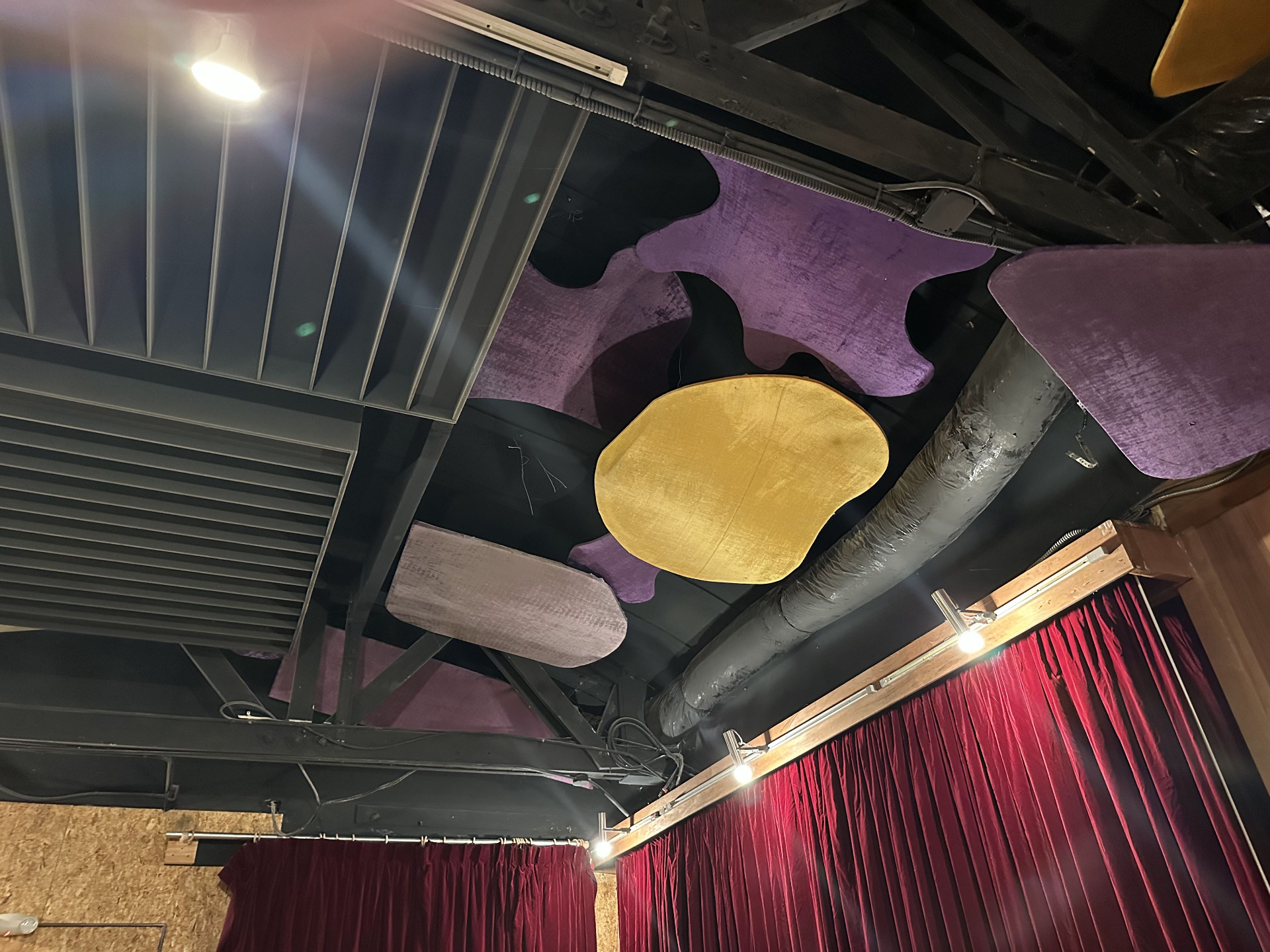 Looking up at the Studio B cloud ceiling — purple velvet amoeba-shaped panels with a yellow oval panel, set between black steel beams and exposed ductwork, with red velvet curtains below.