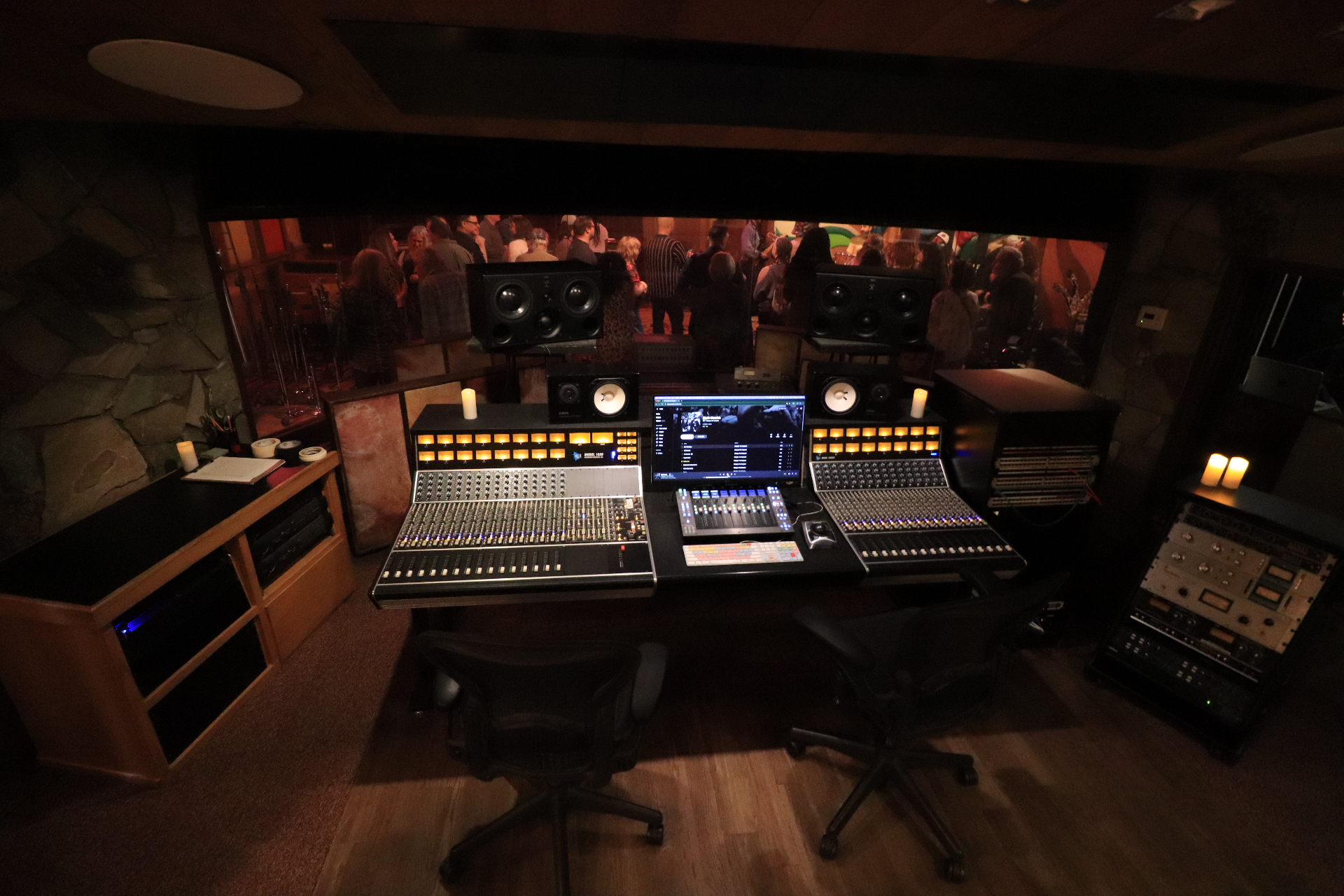 The Studio B control room in 2026 — API console flanked by candles, glowing channel strips, and a view through the glass into the live room during a party.