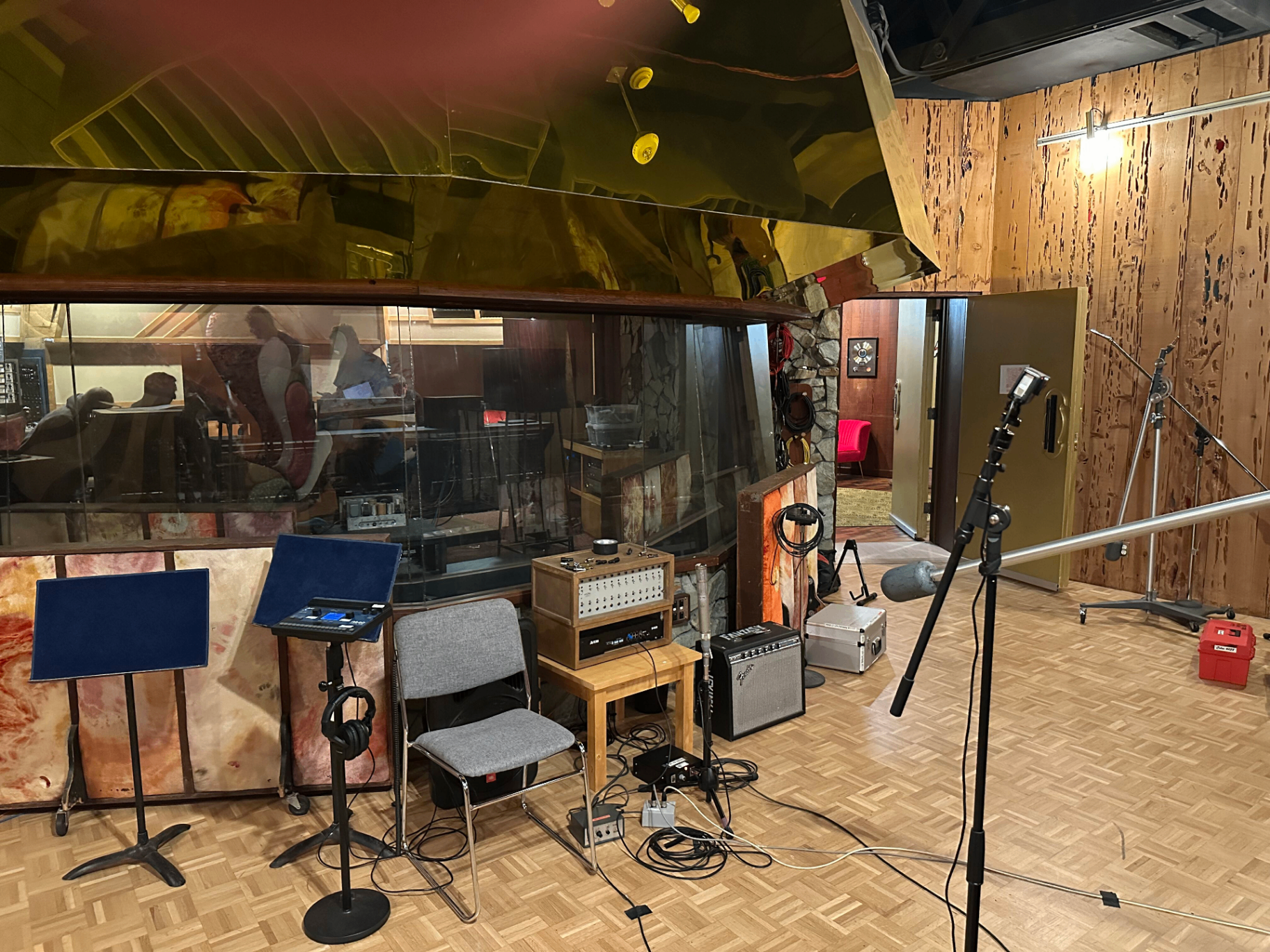 Studio B live room — the curved mirrored ceiling reflecting the control room window, with mic stands, a chair, and amps set up on the parquet floor against the burl-wood wall.