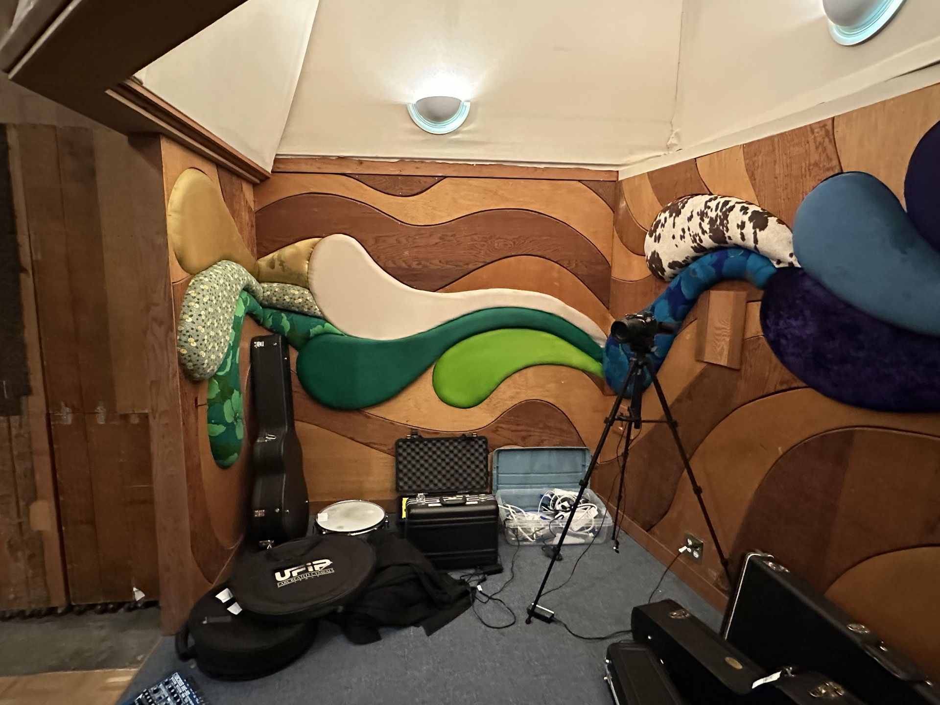 A corner of Studio B with wave-shaped acoustic panels in green sequins, white, lime, blue floral, and cowhide print mounted across a curved redwood wall, with guitar cases and gear on the floor.