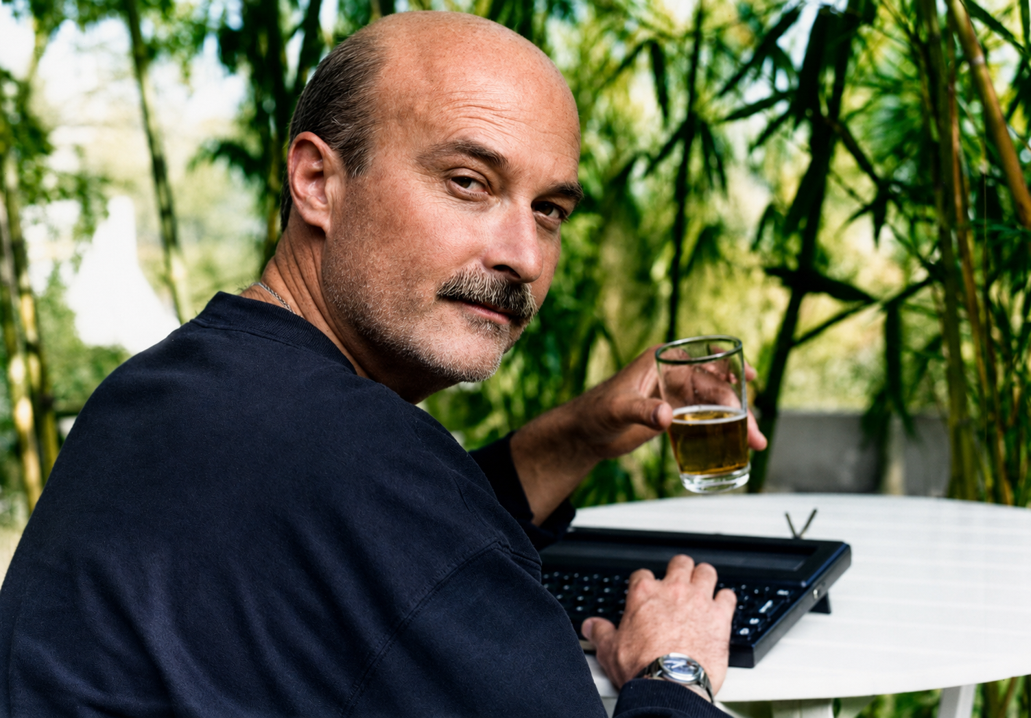 David Goggin in a bamboo garden, holding a whisky glass with a portable keyboard on the table